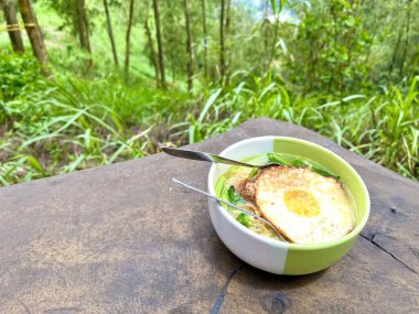 Ahşap masada yumurtalı ramen eriştesi ve arka planda doğa ormanı manzarası.