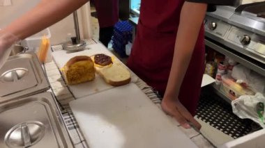 Kırmızı önlüklü bir erkek işçi mutfakta yumurta, sos ve sebzeli biftekli sandviç hazırlıyor..