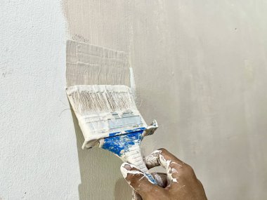 Duvara beyaz boya uygulayan bir fırça tutan, boyama işlemine yakın çekim yapan ve ev dekorasyonu kavramını geliştiren bir el fırçası. Yenileme temaları ve DIY görselleri için ideal.