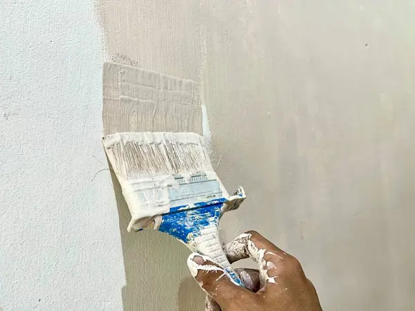 Duvara beyaz boya uygulayan bir fırça tutan, boyama işlemine yakın çekim yapan ve ev dekorasyonu kavramını geliştiren bir el fırçası. Yenileme temaları ve DIY görselleri için ideal.