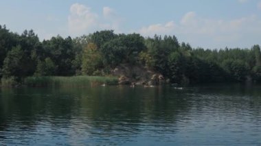 Yazın güzel bir göl. Uzaktaki insanlar gölde yüzüyorlar. Tüm sahil yeşil yapraklı ağaçlarla kaplı. Güzel taş plaj. Bir yaz gününde nehir.