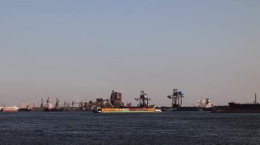 Gemiler Kuzey Denizi boyunca yüzer. Arka planda yel değirmenleri var. Ağır çekim. Hollanda 'daki deniz limanı. Malzeme yüklemek için bir sürü vinç. Farklı gemiler ve mavnalar..