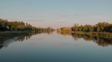 Güzel bir yaz nehri manzarası. Mavi, mavi gökyüzü gölün su yüzeyine yansıyor. Batan güneş yeşil yapraklarla kaplı ağaçları aydınlatıyor. Kıyı boyunca yeşil sazlıklar.