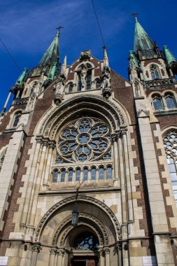 Karmaşık taş oymaları, sivri kemerler ve parlak mavi gökyüzüne karşı kurulmuş büyük bir gül penceresinin yer aldığı neo-gotik bir katedralin yakın görüntüsü..