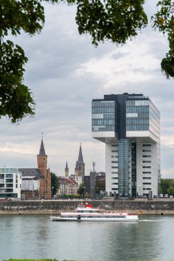 Koln, Kuzey Ren-Vestfalya Germanya - 5.05.2023: Önünde bir tekne bulunan nehir kenarındaki modern cam kuleler. Arka planda ağaç tarafından çerçevelenmiş tarihi kilise kuleleri görülüyor.