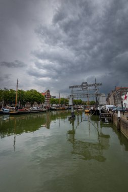 Dramatik bulutlu bir gökyüzünün altında demirli tekneleri olan huzurlu bir liman. İlginç tarihi binalar ve ağaçlar, sakin yeşil suları yansıtarak sakin bir manzara yaratıyor. Dordrecht manzarası.