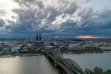 Köln, Almanya 'nın manzarası, ikonik Köln Katedrali, Hohenzollern Köprüsü, Ren Nehri, ve ufuk çizgisinde gün batımına dair ipuçları olan dramatik bulutlu bir gökyüzü..