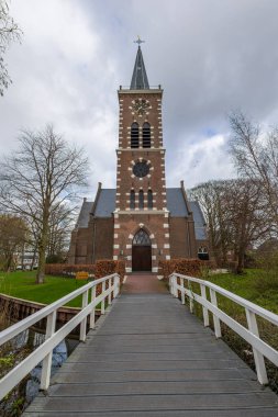 Ağaçlarla çevrili, ahşap bir yaya köprüsünden izlenen, yüksek kulesi olan pitoresk tuğla bir kilise. Bulutlu gökyüzü bu dingin ve zamansız manzaraya derinlik katıyor. Amsterdam yakınlarındaki kilise.