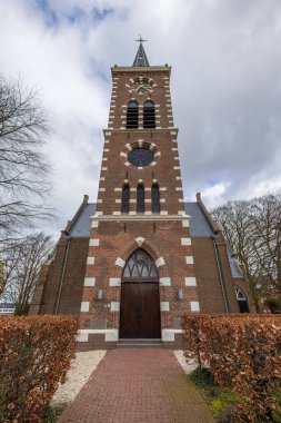 Görünür köklere sahip uzun ve çıplak ağaçlarla çevrili tarihi bir tuğla kilise. Gotik mimari bulutlu bir gökyüzüne vurgulanarak sakin bir atmosfer yaratıyor. Amsterdam yakınlarındaki kilise.
