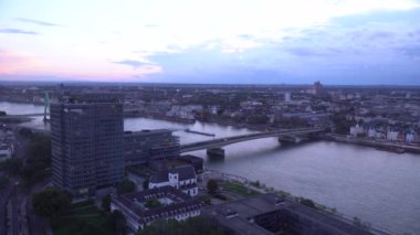 Nehir kenarındaki modern bir şehrin manzarası, bir köprü, yüksek binalar ve tarihi mimari içeriyor. Gökyüzü alacakaranlıkta pastel renklerle hafifçe parlar. Cityscape of Cologne Yukarıdan.