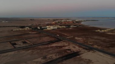 Gün batımında kıyı manzarası, uçsuz bucaksız kumlu düzlükleri kısmen inşa edilmiş binaları, yolları ve ufka karışan sakin bir denizi gösteriyor..
