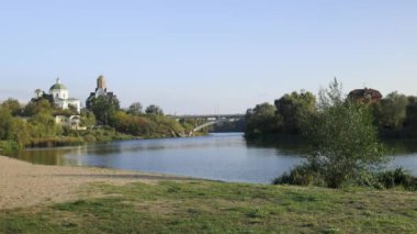 Bereketli bir nehir kenarı manzarası, bir köprü ve tarihi binalar, içinde kubbesi olan bir kilise de var. Sakin su güneşli günün sakin atmosferini yansıtır..