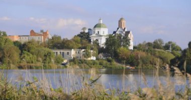 Bereketli bir nehir kenarı manzarası, bir köprü ve tarihi binalar, içinde kubbesi olan bir kilise de var. Sakin su güneşli günün sakin atmosferini yansıtır..