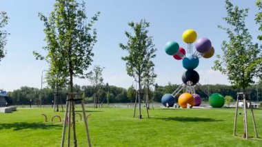 Parlak yeşil çimenler, ağaçlar ve birbirine bağlı büyük kürelerin renkli modern bir heykeli olan güneşli bir park. Arka planda parlak mavi gökyüzü olan huzurlu bir nehir kenarı..