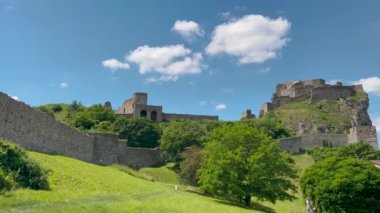Tarihi bir taş kale yeşil bir tepenin tepesinde, parlak mavi bir gökyüzünün altında pofuduk beyaz bulutlarla çevrili, resim gibi ve huzurlu bir ortaçağ manzarası yaratıyor..