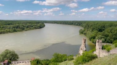 Bereketli mavi gökyüzünün altında tüylü bulutlarla çevrili, bir yamaçta tünemiş antik şato kalıntıları olan sakin bir nehir kenarı manzarası. Tarihle doğanın mükemmel bir karışımı..