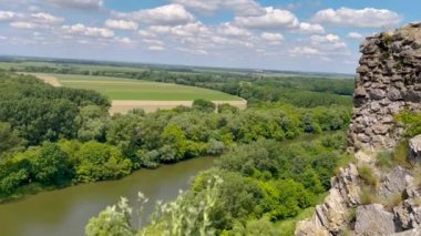 Bereketli mavi gökyüzünün altında tüylü bulutlarla çevrili, bir yamaçta tünemiş antik şato kalıntıları olan sakin bir nehir kenarı manzarası. Tarihle doğanın mükemmel bir karışımı..