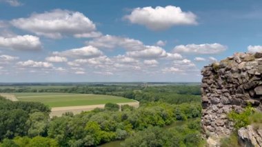 Bereketli mavi gökyüzünün altında tüylü bulutlarla çevrili, bir yamaçta tünemiş antik şato kalıntıları olan sakin bir nehir kenarı manzarası. Tarihle doğanın mükemmel bir karışımı..