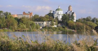 Bereketli bir nehir kenarı manzarası, bir köprü ve tarihi binalar, içinde kubbesi olan bir kilise de var. Sakin su güneşli günün sakin atmosferini yansıtır..