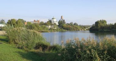 Bereketli bir nehir kenarı manzarası, bir köprü ve tarihi binalar, içinde kubbesi olan bir kilise de var. Sakin su güneşli günün sakin atmosferini yansıtır..