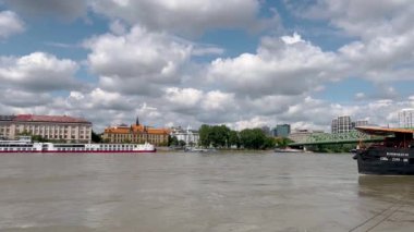 Büyük bir yolcu gemisi, tarihi Avrupa mimarisi, yeşil bir köprü ve durgun sulara yansıyan tüylü beyaz bulutlarla dolu parlak bir gökyüzü içeren huzurlu bir nehir manzarası..