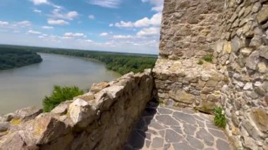 Eski bir kalenin taş duvarlarından çarpıcı bir nehir manzarası. Durgun nehir, parlak mavi gökyüzünün altında yemyeşil ormanlar arasında akar. Doğa ve tarihi uyum içinde sergiler..