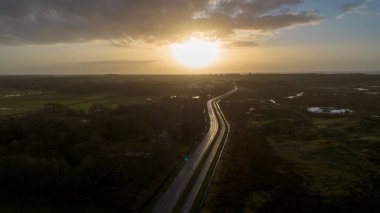 Altın saat boyunca çekilen bir video, kırsal bir arazide, su yansıtmalı tarlalarda, ve uzak binalarda, sıcak, günbatımı aydınlığında bir gökyüzünün altında, dolambaçlı bir yolu gösteriyor..