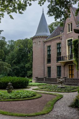 Etrafı yemyeşil, manikürlü bahçeler ve huzurlu bulutlu gökyüzü ile çevrili yuvarlak bir kulesi, sivri çatılı ve kemerli pencereli tarihi tuğla bir malikâne manzarası.