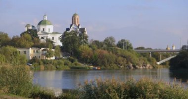 Bereketli bir nehir kenarı manzarası, bir köprü ve tarihi binalar, içinde kubbesi olan bir kilise de var. Sakin su güneşli günün sakin atmosferini yansıtır..