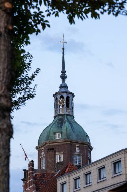 Yeşil kubbeli tarihi bir saat kulesi ve tuğla ve modern binaların üzerinde yükselen bir kule. Akşam gökyüzü ve ağaç dalları sahneyi çerçeveleyerek Avrupa mimari düzenine derinlik katıyor.