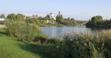 Bereketli bir nehir kenarı manzarası, bir köprü ve tarihi binalar, içinde kubbesi olan bir kilise de var. Sakin su güneşli günün sakin atmosferini yansıtır..