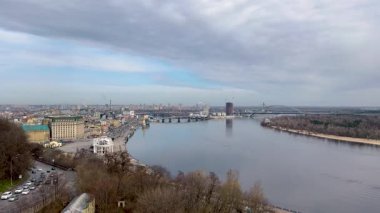 Nehir, köprüler ve modern binaları olan panoramik bir şehir manzarası. Gökyüzü parçalı bulutlu ve yol boyunca trafik hareket ediyor. Sakin nehir ufku yansıtıyor, şehir manzarası yaratıyor..