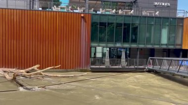 Bratislava, Bratislava Bölgesi, Slovak Cumhuriyeti - 09.07.2024: Hızla akan bir nehrin üzerinde ahşap panelli modern bir binaya karşı yüzen ağaç dalları yığılıyor. Küçük metal bir köprü birleşir.
