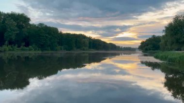 Sakin bir göl, yemyeşil ağaçlarla çevrili günbatımının altın tonlarını yansıtır. Suda hafif dalgalanmalar oluşur ve gökyüzü yumuşak maviler ve ılık portakalların karışımıdır. Huzurlu bir manzara yaratır.