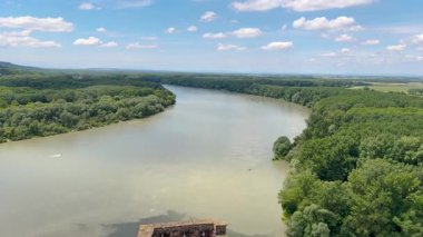 Bereketli yeşillikler, yuvarlanan tepeler ve parlak, güneşli bir gökyüzünün altında ön planda taş bir duvarla çevrili geniş bir nehrin manzarası..