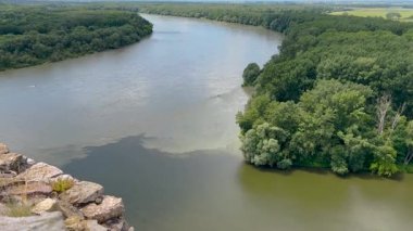 Bereketli yeşillikler, yuvarlanan tepeler ve parlak, güneşli bir gökyüzünün altında ön planda taş bir duvarla çevrili geniş bir nehrin manzarası..