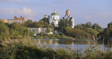 Bereketli bir nehir kenarı manzarası, bir köprü ve tarihi binalar, içinde kubbesi olan bir kilise de var. Sakin su güneşli günün sakin atmosferini yansıtır..