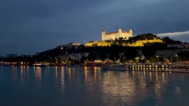 Bratislava Kalesi 'nin manzaralı bir gece manzarası, Tuna Nehri' ne bakan bir tepenin tepesinde aydınlandı. Şehir ışıklarının yansımaları, nehir kıyısı boyunca yanaşmış teknelerle suda parıldıyor..