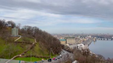 Nehir, köprüler ve modern binaları olan panoramik bir şehir manzarası. Gökyüzü parçalı bulutlu ve yol boyunca trafik hareket ediyor. Sakin nehir ufku yansıtıyor, şehir manzarası yaratıyor..