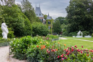 Etrafı yemyeşil, manikürlü bahçeler ve huzurlu bulutlu gökyüzü ile çevrili yuvarlak bir kulesi, sivri çatılı ve kemerli pencereli tarihi tuğla bir malikâne manzarası.