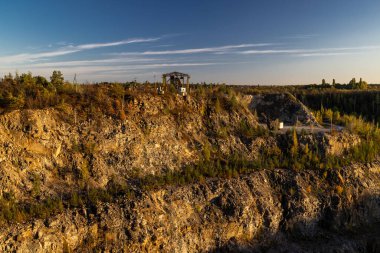 Kayalık bir uçurum, seyrek ağaçlar ve sıcak güneş ışığı altında kuru çalılar. Tepede, terk edilmiş bir endüstriyel yapı sonbahar yaprakları arasında duruyor ve doğa ile insan yapımı kalıntılar arasında bir zıtlık oluşturuyor..
