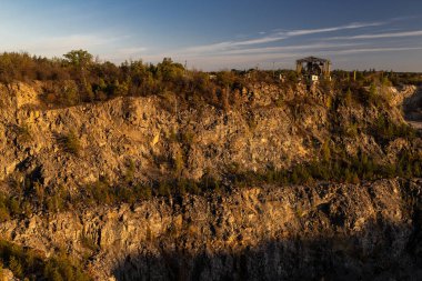 Kayalık bir uçurum, seyrek ağaçlar ve sıcak güneş ışığı altında kuru çalılar. Tepede, terk edilmiş bir endüstriyel yapı sonbahar yaprakları arasında duruyor ve doğa ile insan yapımı kalıntılar arasında bir zıtlık oluşturuyor..