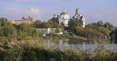 Bereketli bir nehir kenarı manzarası, bir köprü ve tarihi binalar, içinde kubbesi olan bir kilise de var. Sakin su güneşli günün sakin atmosferini yansıtır..