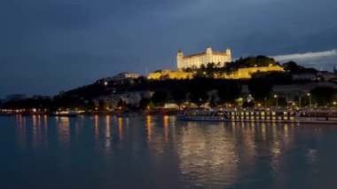Bratislava Kalesi 'nin manzaralı bir gece manzarası, Tuna Nehri' ne bakan bir tepenin tepesinde aydınlandı. Şehir ışıklarının yansımaları, nehir kıyısı boyunca yanaşmış teknelerle suda parıldıyor..