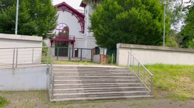 Kırmızı ahşap detaylara, geniş kemerli pencerelere ve yemyeşil ağaçlarla çevrili tarihi beyaz bir bina. Sahne, modern eskrim ve parlak bir gökyüzünün altında bir lamba direği tarafından çerçevelenmiştir.