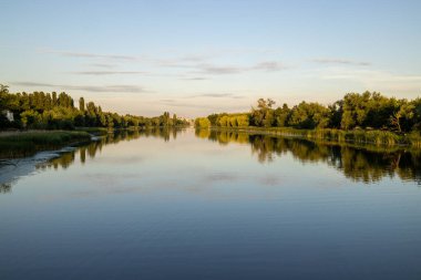 Bereketli yeşil ağaçları ve altın bir akşam gökyüzünü yansıtan sakin bir nehir. Durgun su, sakin manzarayı yansıtıyor. Gün batımında huzurlu ve resimsel bir doğa manzarası yaratıyor..