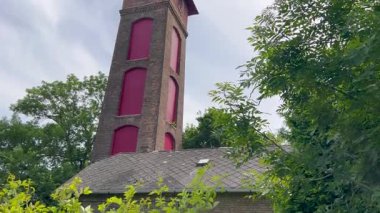 Kırmızı pencere kaplamalı uzun bir tuğla kule, yemyeşil ağaçlarla çevrili parçalı bulutlu bir gökyüzüne karşı duruyor. Sahne, mimariyle doğayı harmanlayan tarihi ve gizemli bir atmosfere sahiptir.