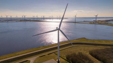 Büyük bir rüzgar türbini, güneş ışığını yansıtan kıyı sularının yanında duruyor. Ufukta daha fazla türbin var, yenilenebilir enerji üretiyor. Kavisli yollar ve yeşil alanlar bu manzarayı tamamlıyor.