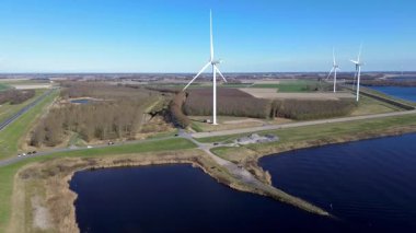 Suyun üzerinde birden fazla rüzgar türbini bulunan kıyı rüzgâr çiftliğinin manzaralı bir görüntüsü. Virajlı yollar manzaranın içinden geçiyor, arabalar görünür durumda. Güneş sudan yansıyor..