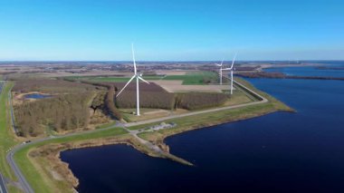 Suyun üzerinde birden fazla rüzgar türbini bulunan kıyı rüzgâr çiftliğinin manzaralı bir görüntüsü. Virajlı yollar manzaranın içinden geçiyor, arabalar görünür durumda. Güneş sudan yansıyor..